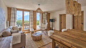 A spacious living area with a wooden dining table, a sofa, and a chair. There are large windows with curtains, a hanging light fixture, and a view of the outdoors.
