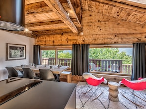 Un coin repas en bois avec une table et des chaises, une grande fenêtre avec des rideaux et une vue sur la verdure à l’extérieur.