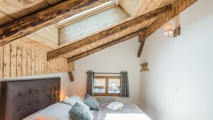 A bedroom with a wooden ceiling, a large bed with pillows and a blanket, a window with curtains, and a wall-mounted lamp.