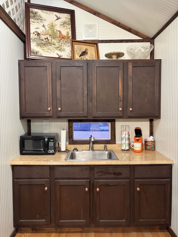 Una cocina con gabinetes de madera oscura, un lavamanos de acero inoxidable, un microondas y una ventana sobre el lavamanos.