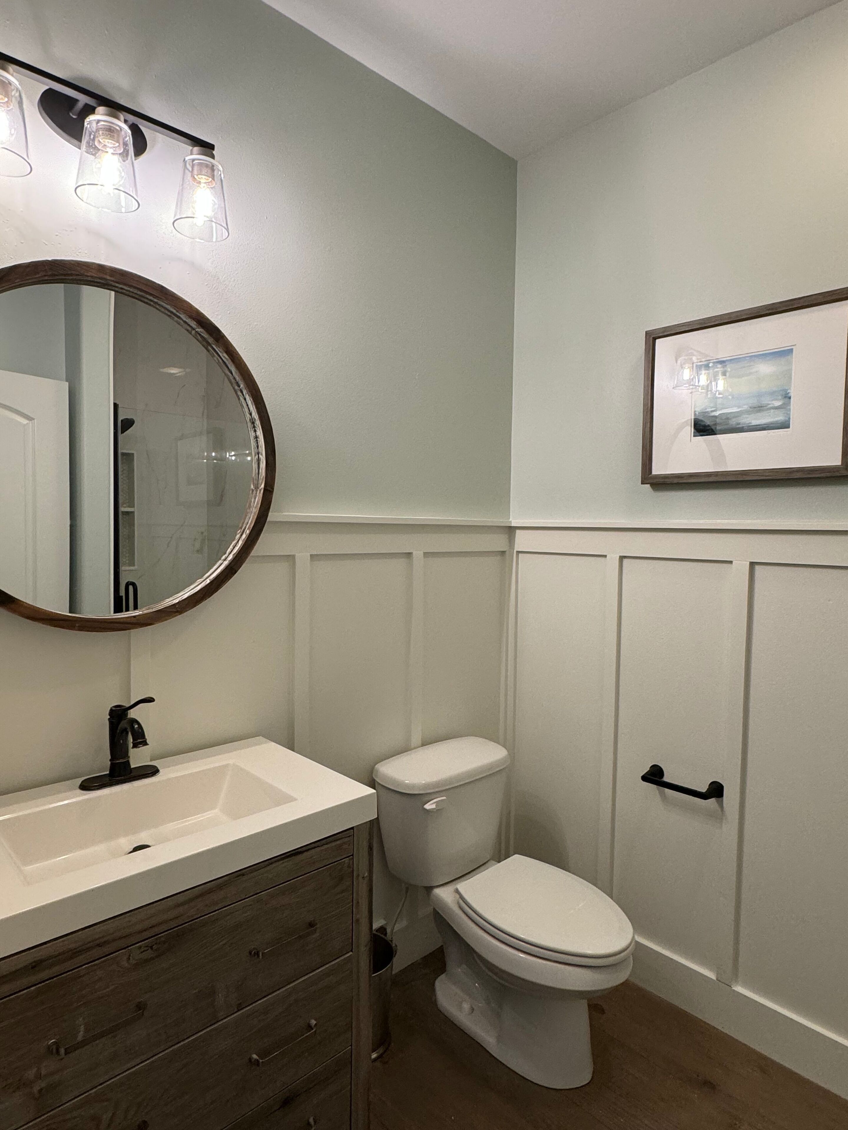 A bathroom with a white vanity, a round mirror, a toilet, and a framed picture on the wall.