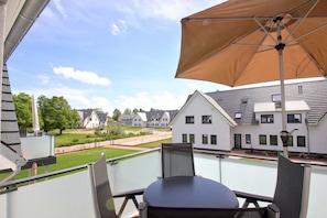 A balcony with a table and chairs, a large umbrella, and a view of a residential area with houses and a lawn.
