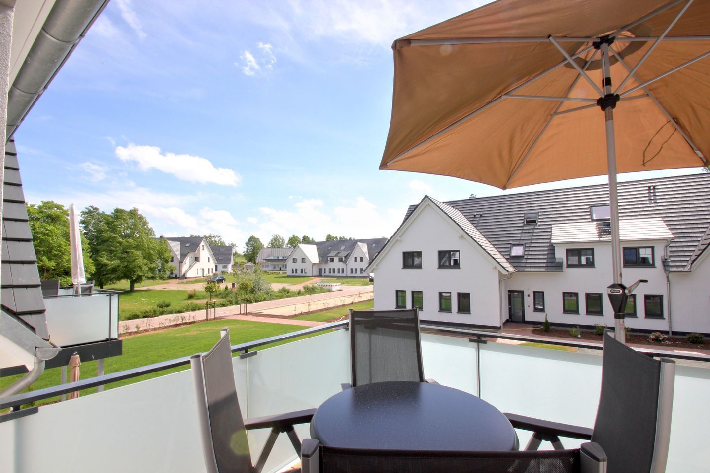 Een balkon met een tafel en stoelen, een grote parasol en uitzicht op een woonwijk met huizen en een gazon.