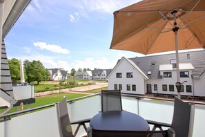 A balcony with a table and chairs, a large umbrella, and a view of a residential area with houses and a lawn.