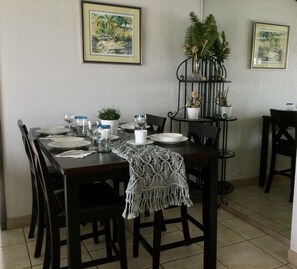 A dining area with a table set for four, surrounded by chairs. There are framed pictures on the wall, a potted plant on a stand, and a macrame table runner. - Beachside, Pool, Amazing Views, Excellent Location - 3bedroom/3bathroom Condo (St Thomas)