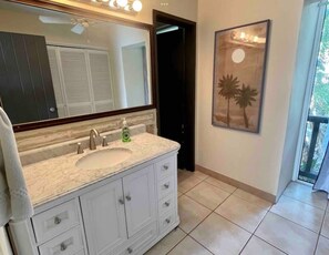A bathroom with a large mirror, a sink, and a cabinet. There is a door leading to another room and a window with a view of palm trees. - Beachside, Pool, Amazing Views, Excellent Location - 3bedroom/3bathroom Condo (St Thomas)