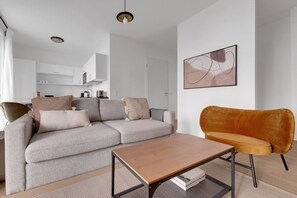 A modern living room with a grey sectional sofa, a wooden coffee table, and a mustard-colored armchair. A kitchen is visible in the background.