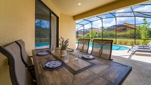 An outdoor dining area with a table set for four, surrounded by chairs, and a view of a swimming pool and greenery. - Providence Golf & Country Club Pool Home in Gated & Guarded Community (Davenport)