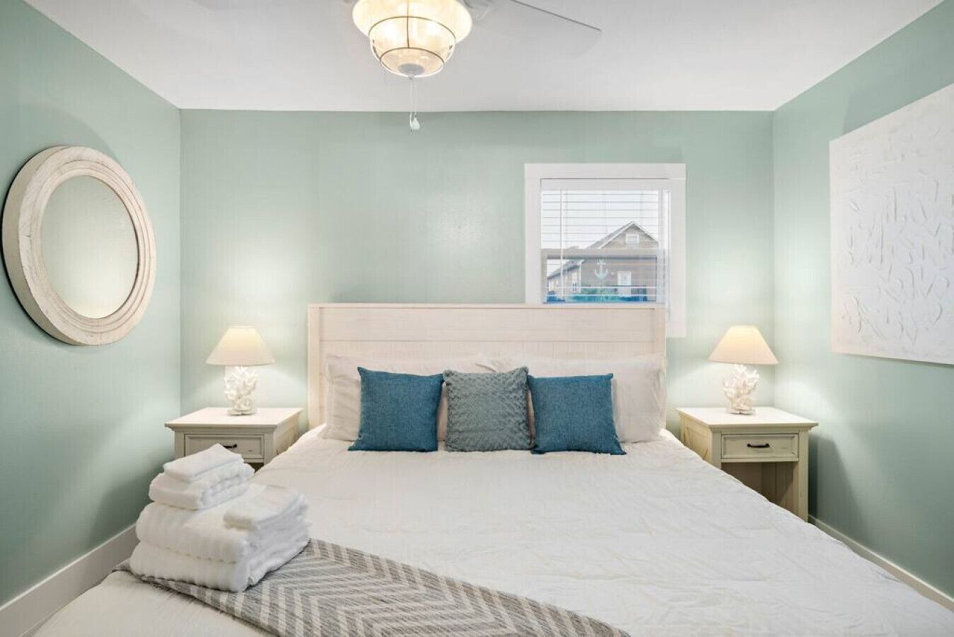 A bedroom with a bed, two bedside tables, a round mirror, a window with a view, and a ceiling fan.