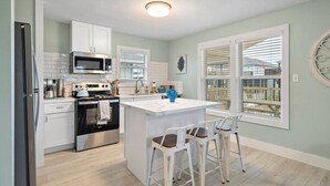 A modern kitchen with white cabinetry, a central island with bar stools, a stainless steel refrigerator, and a microwave. - Canalfront Home with Huge Deck, Close to the Beach (Galveston)