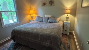 A bedroom with a large bed, two bedside tables with lamps, a window with a view of trees, and two wall art pieces. - Hidden Gem 10 mins to Downtown or Wrightsville Beach. Brand New King Sized Bed (Wilmington)