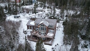A large, multi-story house with a prominent front porch and multiple decks, surrounded by a snowy landscape and dense forest.