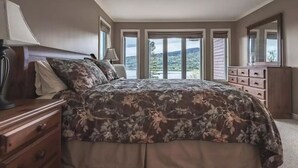 A bedroom with a large bed, a dresser, a lamp, and a view of the outdoors.
