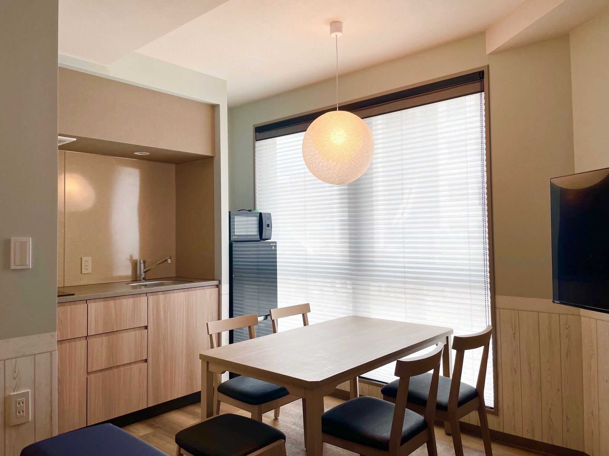 A modern kitchen with a dining area, wooden cabinets, a built-in microwave, and a large window with blinds.