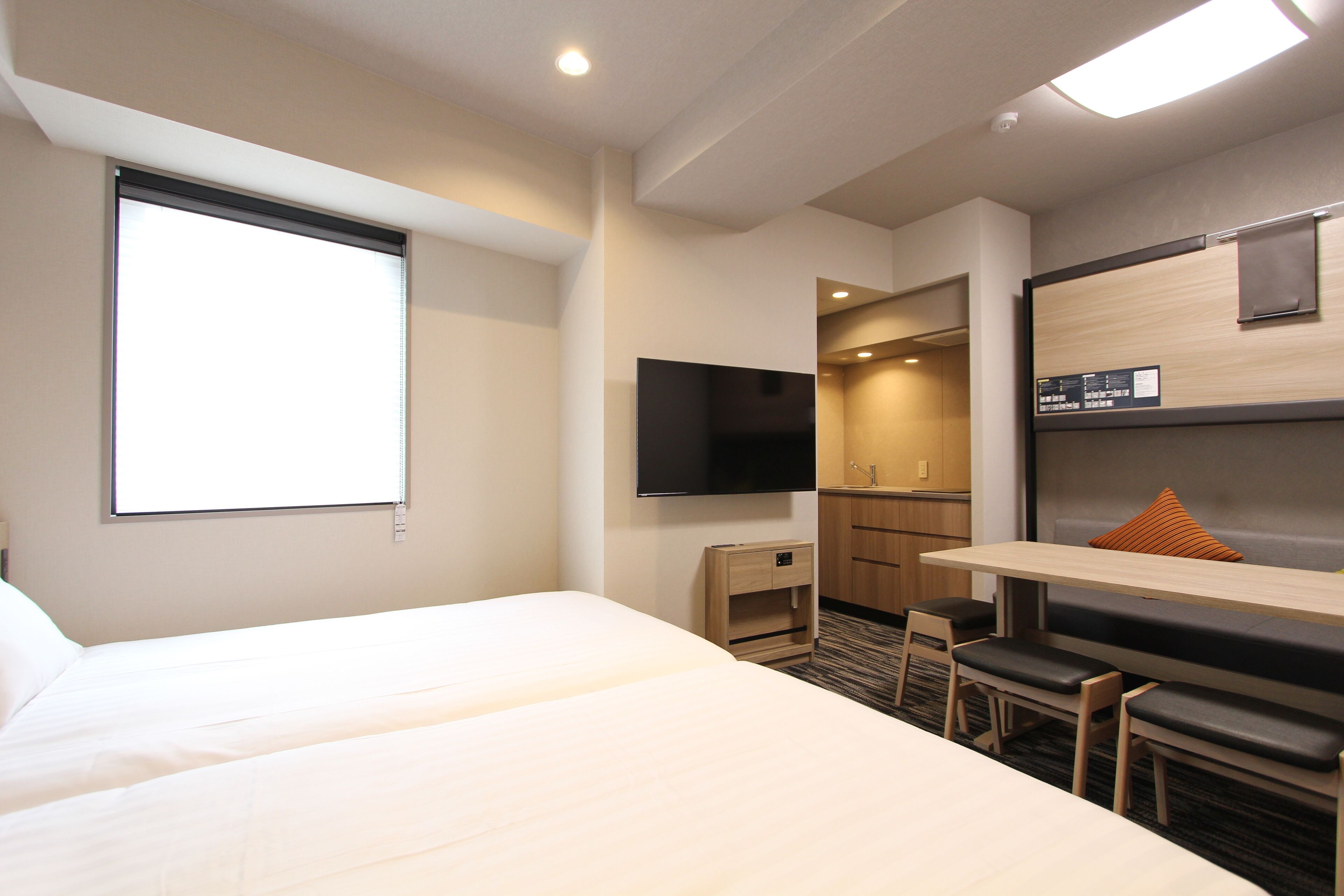 Quarto de hotel com uma cama grande, uma escrivaninha com cadeira, televisão e uma pequena cozinha equipada.