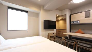 Quarto de hotel com uma cama grande, uma escrivaninha com cadeira, televisão e uma pequena cozinha equipada.