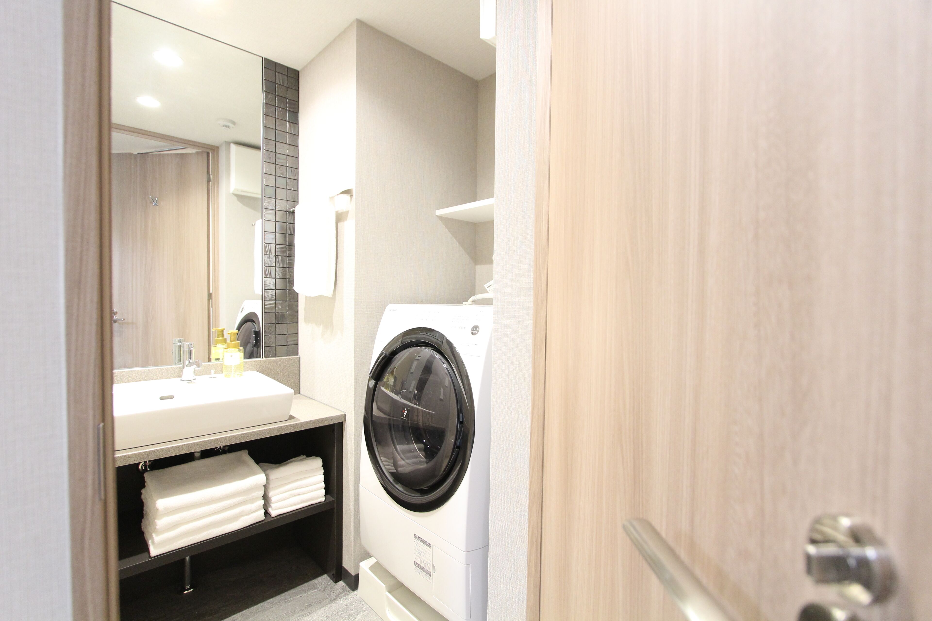 A compact hotel bathroom with a sink, mirror, and a front-loading washing machine.