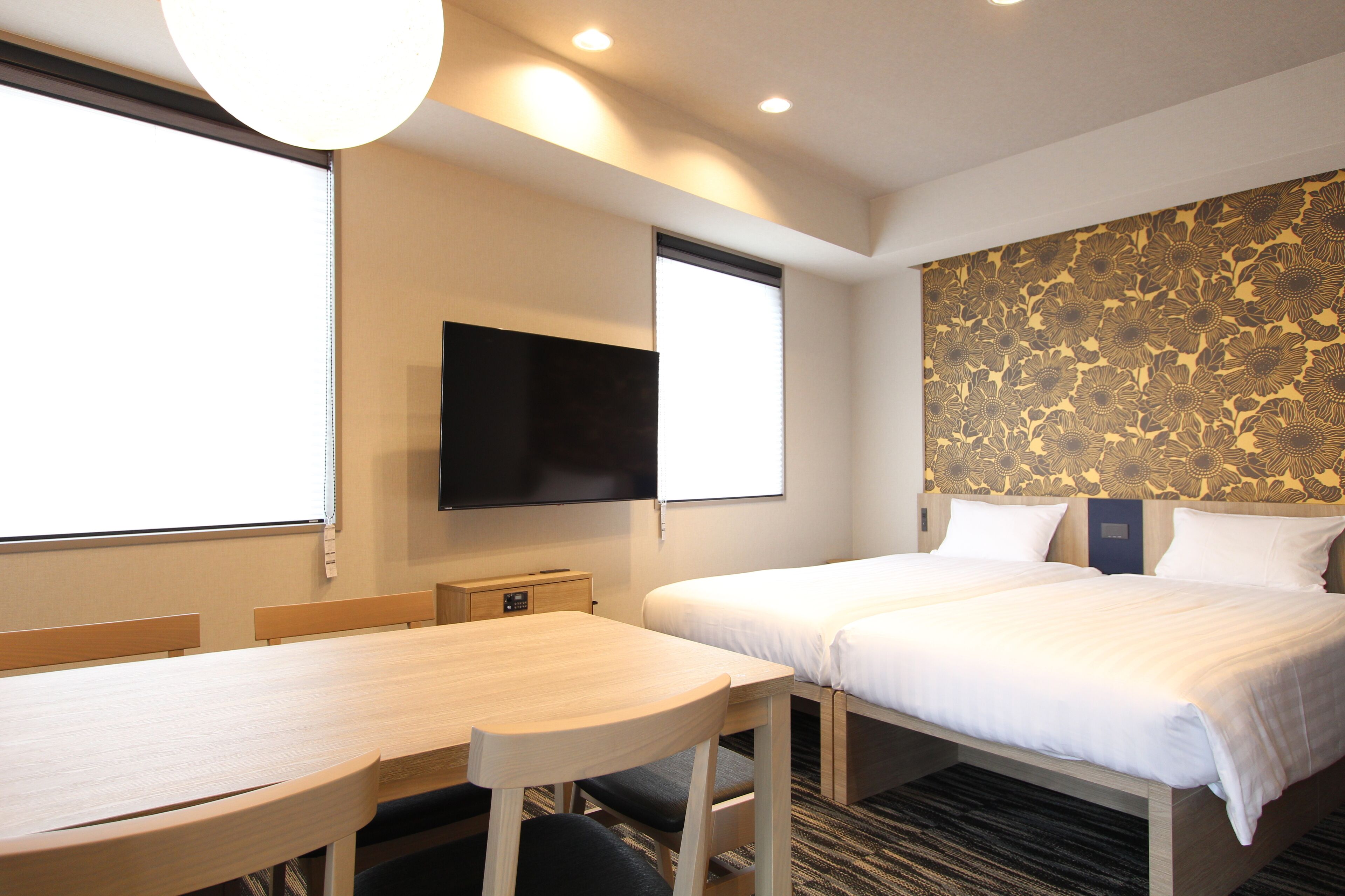 A hotel room with two beds, a flat-screen TV mounted on the wall, a wooden table, and a window with blinds.