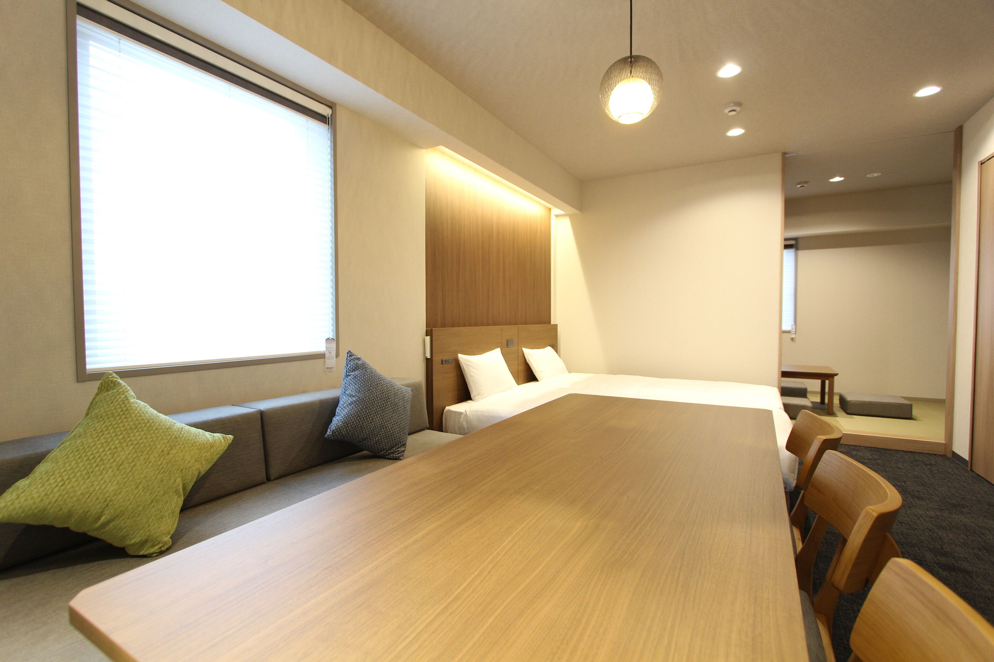 Una habitación de hotel con una amplia mesa de madera, un sofá con cojines, una cama con ropa de cama blanca y una pequeña mesa con un banco.