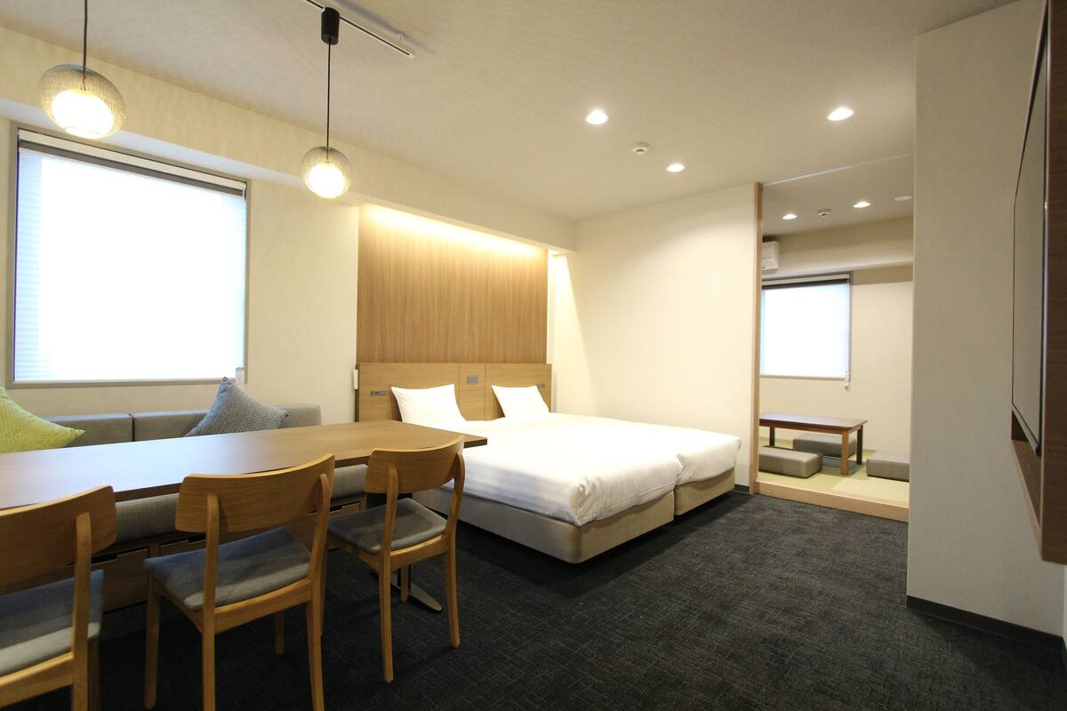 Ein Hotelzimmer mit einem Bett, einem Schreibtisch und Stühlen. Es gibt Fenster mit Jalousien, eine Deckenleuchte und ein an der Wand befestigtes Fernsehgerät.