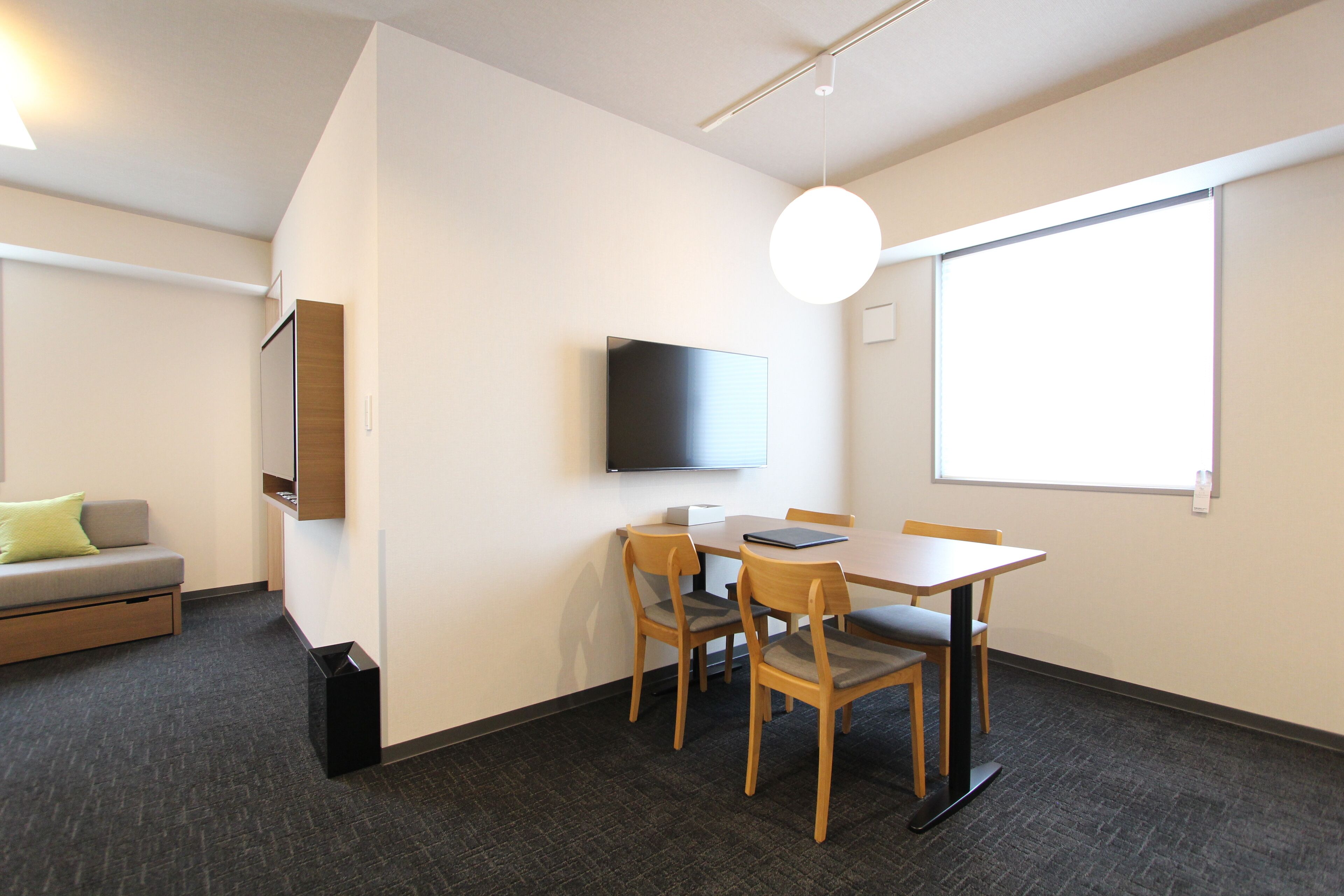 Une chambre moderne dotée d’un téléviseur à écran plat fixé au mur, d’une table en bois avec quatre chaises, d’un canapé gris agrémenté d’un coussin vert et d’une grande fenêtre laissant entrer la lumière naturelle.