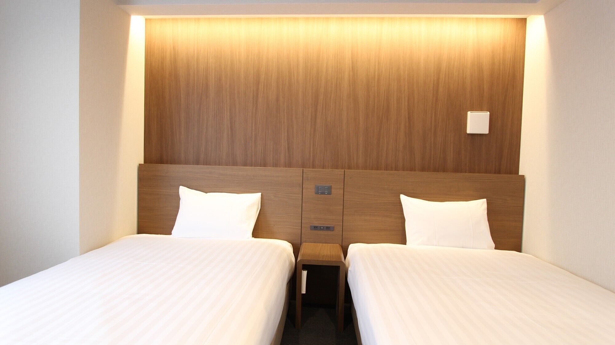Quarto de hotel com duas camas, uma cabeceira de madeira e uma mesa de cabeceira com abajur.