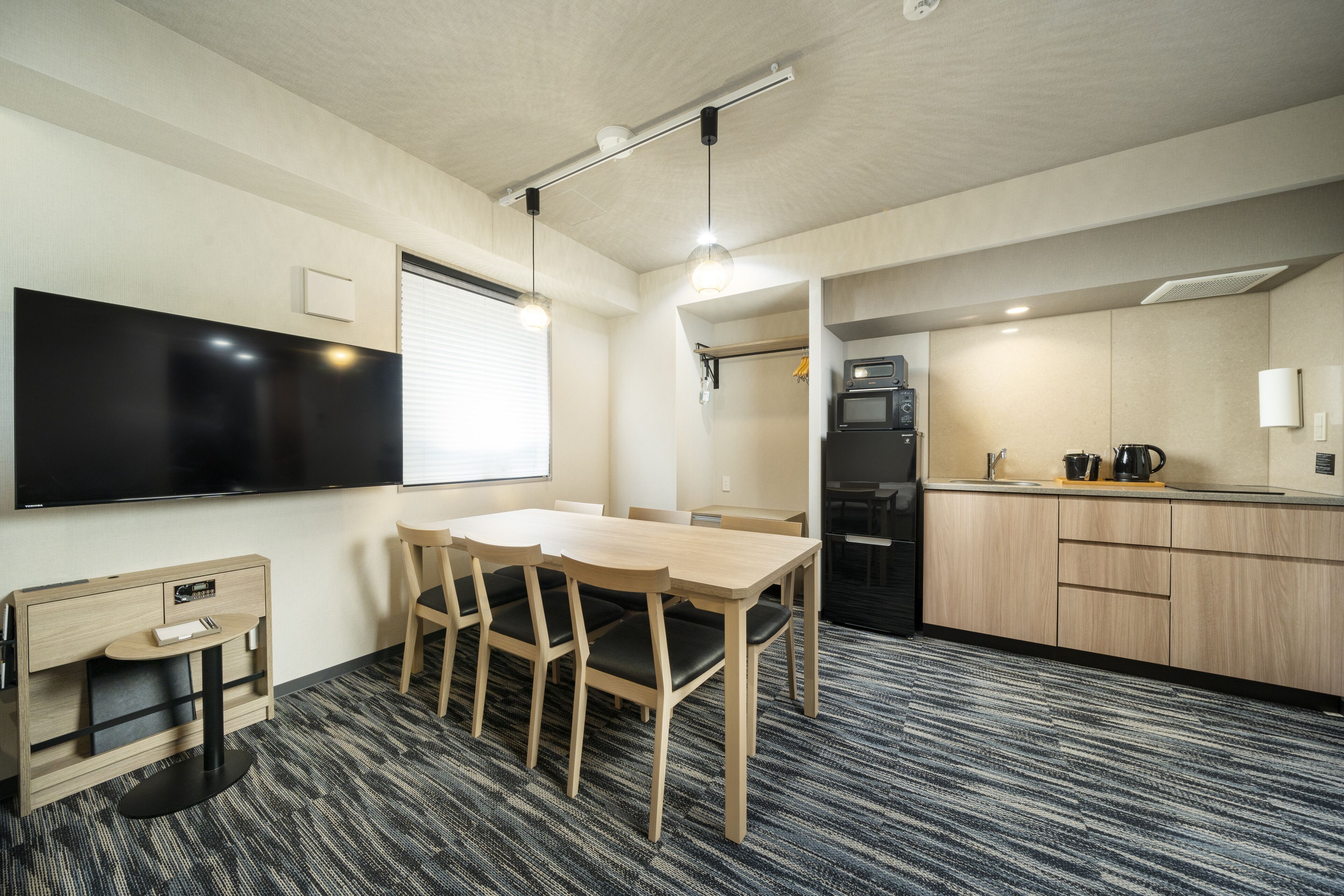 A modern hotel room with a flat-screen TV, a wooden dining table with chairs, a small side table, a kitchenette with a microwave, and a large window with blinds.