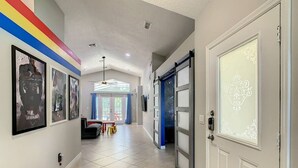 A hallway with a tiled floor, framed artwork on the walls, a door with a decorative glass panel, and a small table with chairs in the distance. - Single Level 5 Bedrm/Pool/SPA/3Miles Disney (Kissimmee)