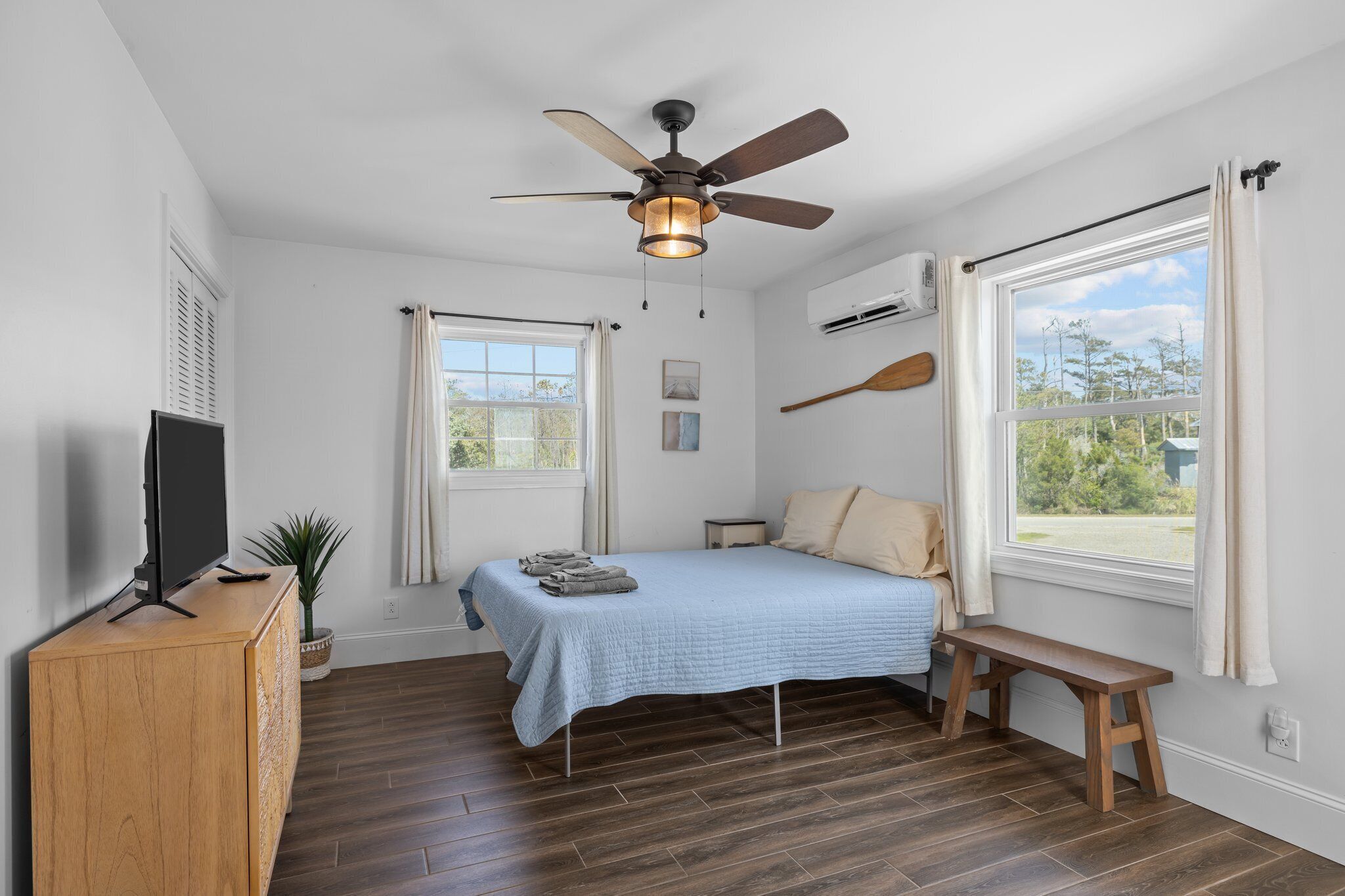 A bedroom with a bed, a ceiling fan, a TV on a wooden cabinet, a window with curtains, and a view of the outdoors.