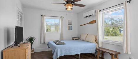 A bedroom with a bed, a ceiling fan, a TV on a wooden cabinet, a window with curtains, and a view of the outdoors.