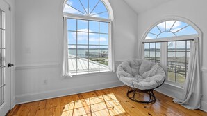 Quarto com piso de madeira, uma cadeira branca e duas grandes janelas em arco com vista para um cais e para a água.
