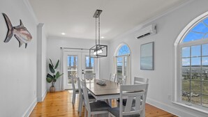 Uma sala de jantar com mesa de madeira e cadeiras brancas, luminária pendente e uma grande janela com vista para o exterior.
