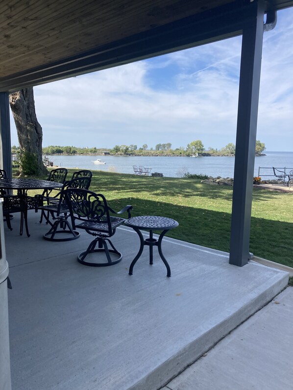 An outdoor patio with metal tables and chairs, overlooking a lake with boats and a grassy area.