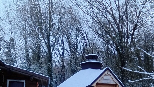 Une cabane en bois dont le toit est recouvert de neige, avec une porte d’entrée dotée d’une fenêtre ronde et un banc à l’extérieur.