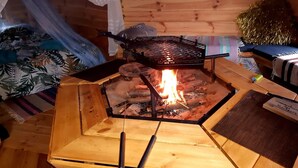 Un barbecue extérieur en bois, avec un feu allumé en dessous, entouré de meubles en bois et d’une plante décorative.