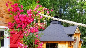 Une cabane en bois avec un toit noir, entourée d’une végétation luxuriante et de fleurs roses.