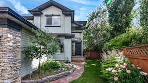 A two-story house with a stone exterior, a brick pathway leading to the front door, and a garden with pink flowers. - G@Home Quiet 5BR4BA Retreat, Gateway to Banff & COP & Hwy 1 (Calgary)