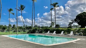 A swimming pool with clear blue water, surrounded by white lounge chairs and palm trees under a partly cloudy sky. - Best View in Hilo - Pool, WIFI, 2 BD Condo (Hilo)