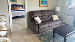 A living room with a brown sofa, a small table, a lamp, and a bed in the background. - Best View in Hilo - Pool, WIFI, 2 BD Condo (Hilo)