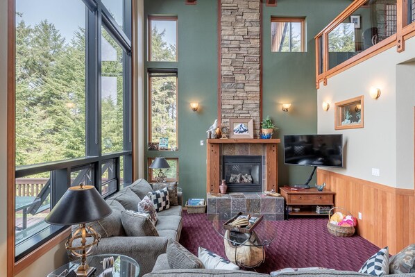 A spacious living room with a stone fireplace, large windows, and a flat-screen TV.