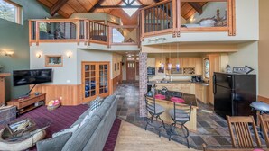 A spacious loft-style living area with a high ceiling, wooden beams, a large TV, a fireplace, and a kitchen with a refrigerator and bar seating.
