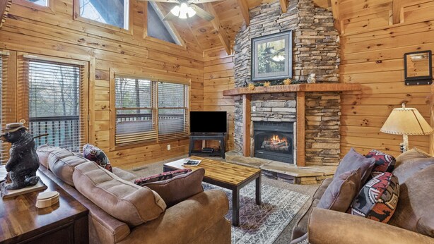 A cozy log cabin interior with a stone fireplace, a television, and a comfortable seating area with a sofa and armchairs.