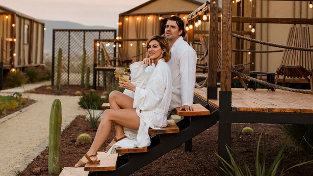 Una pareja sentada en un banco de madera al aire libre, con un paisaje desértico y edificios en el fondo.