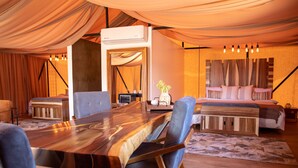 Una habitación tipo tienda de campaña con una mesa de comedor de madera, sillas tapizadas en azul, una cama con ropa de cama estampada y un aire acondicionado de pared.