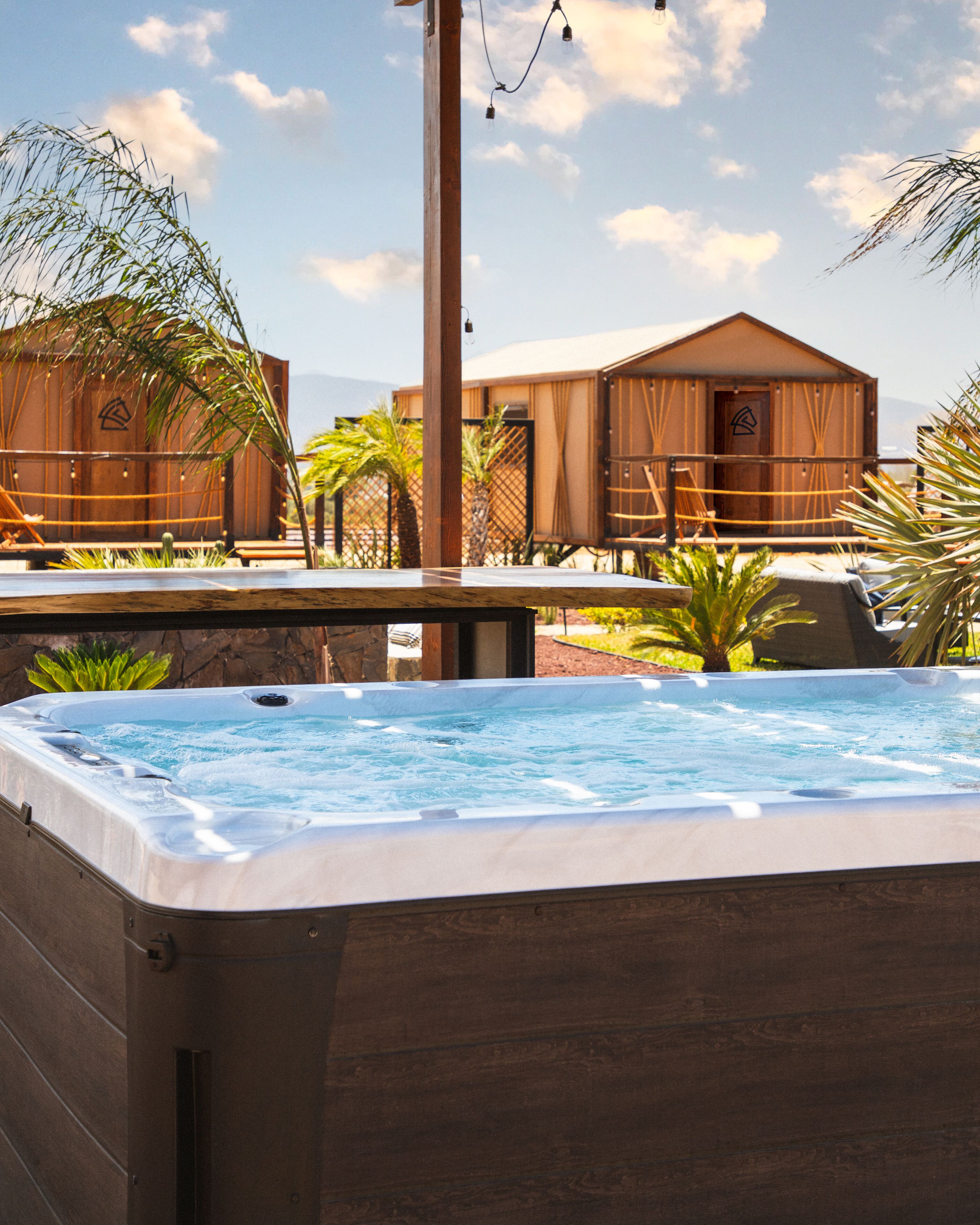 A hot tub with a wooden deck, surrounded by palm trees and a wooden building in the background.