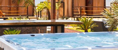 A hot tub with a wooden deck, surrounded by palm trees and a wooden building in the background.