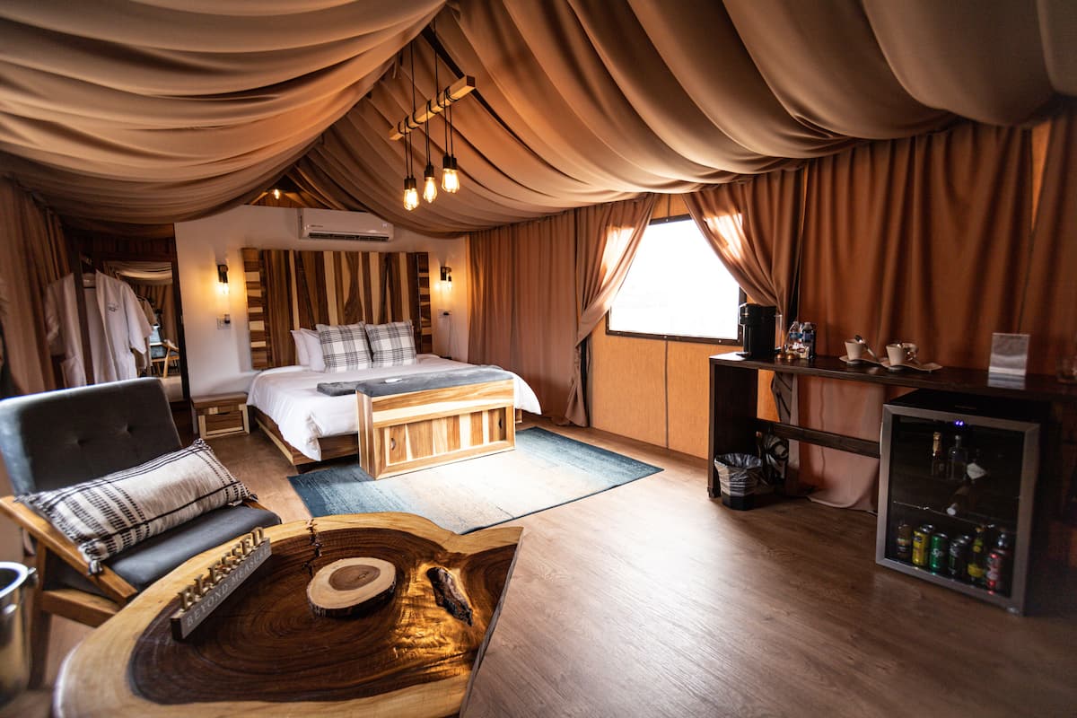 A spacious bedroom with a large bed, a wooden coffee table, a chair, and a minibar.