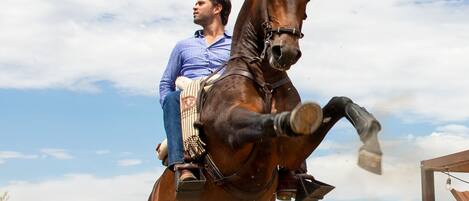 Una persona montando a caballo en un área de establos con cercas de madera y cactus.
