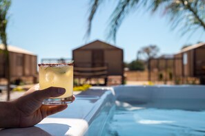 A hand holding a drink near a pool with a clear sky and palm trees in the background. - Indomito Resort & Hotel Boutique (Valle de Guadalupe)