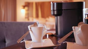 Una preparación de café con una taza y platillo sobre una mesa, una cafetera y un sofá al fondo.